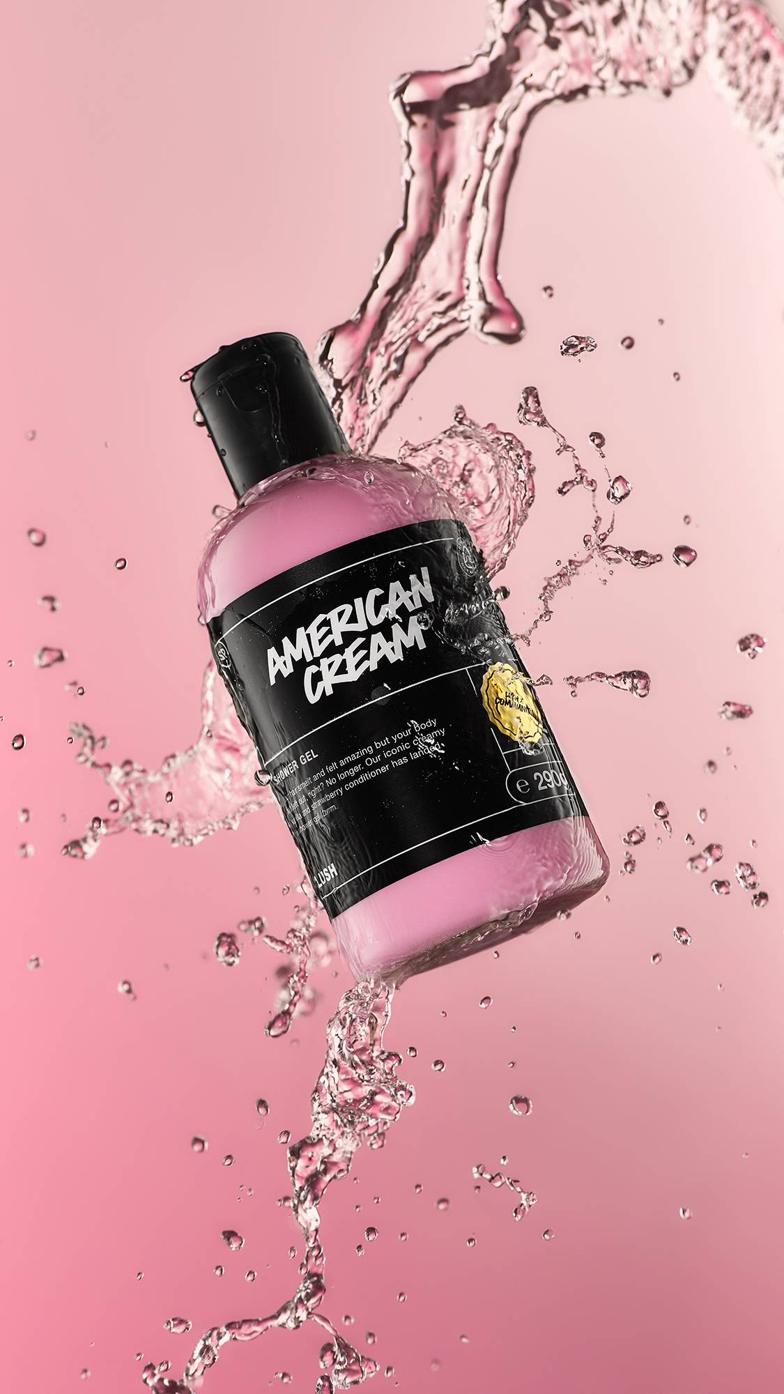 A high-definition action shot of the American Cream shower gel bottle mid-air being splashed by fresh water surrounded by droplets on a candyfloss-pink background. 