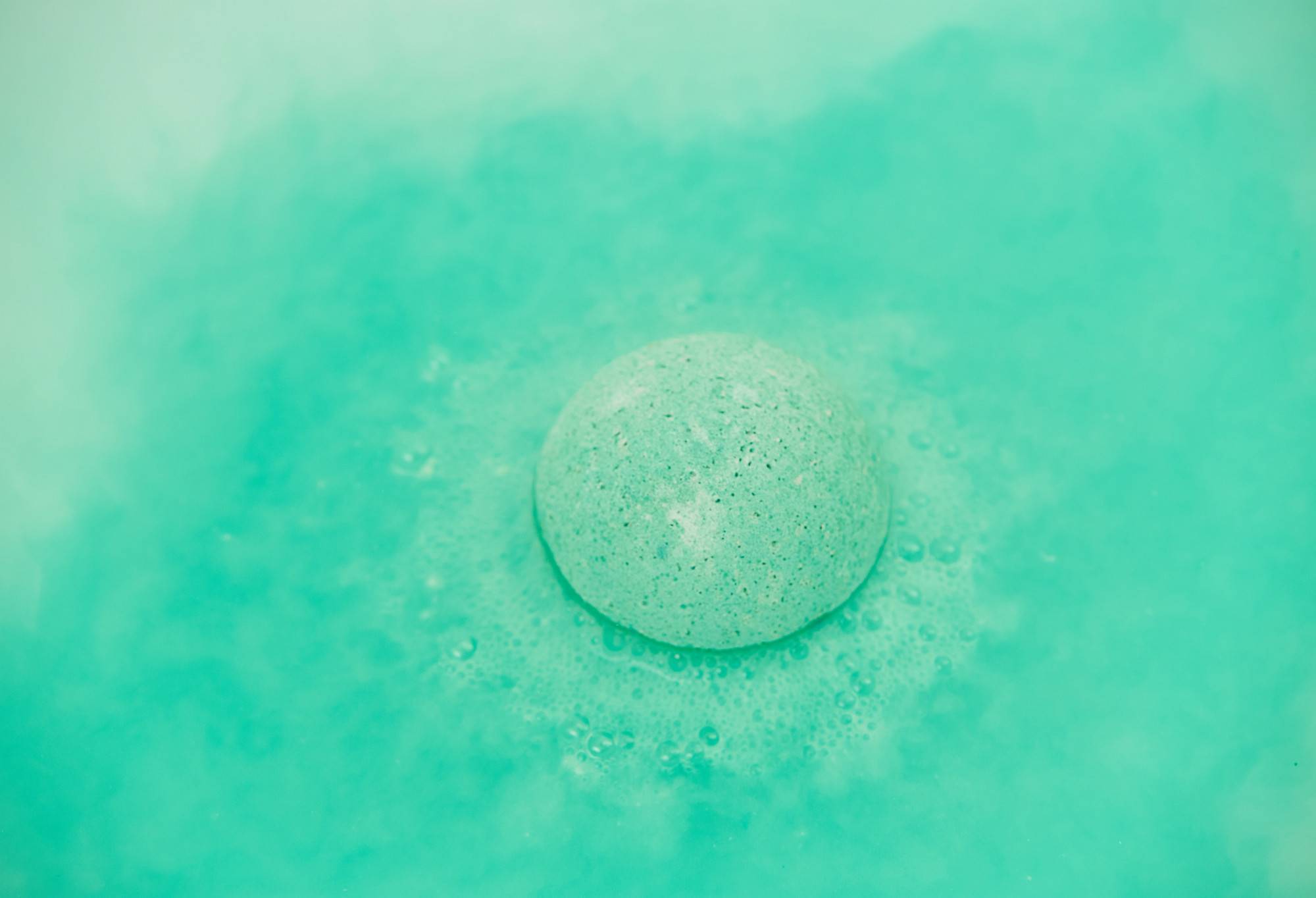 The Avobath bath bomb sits perfectly central, floating on the bathwater surface as it starts to dissolve. There are delicate bubbles being given off and the water is a beautiful, fresh turquoise colour. 