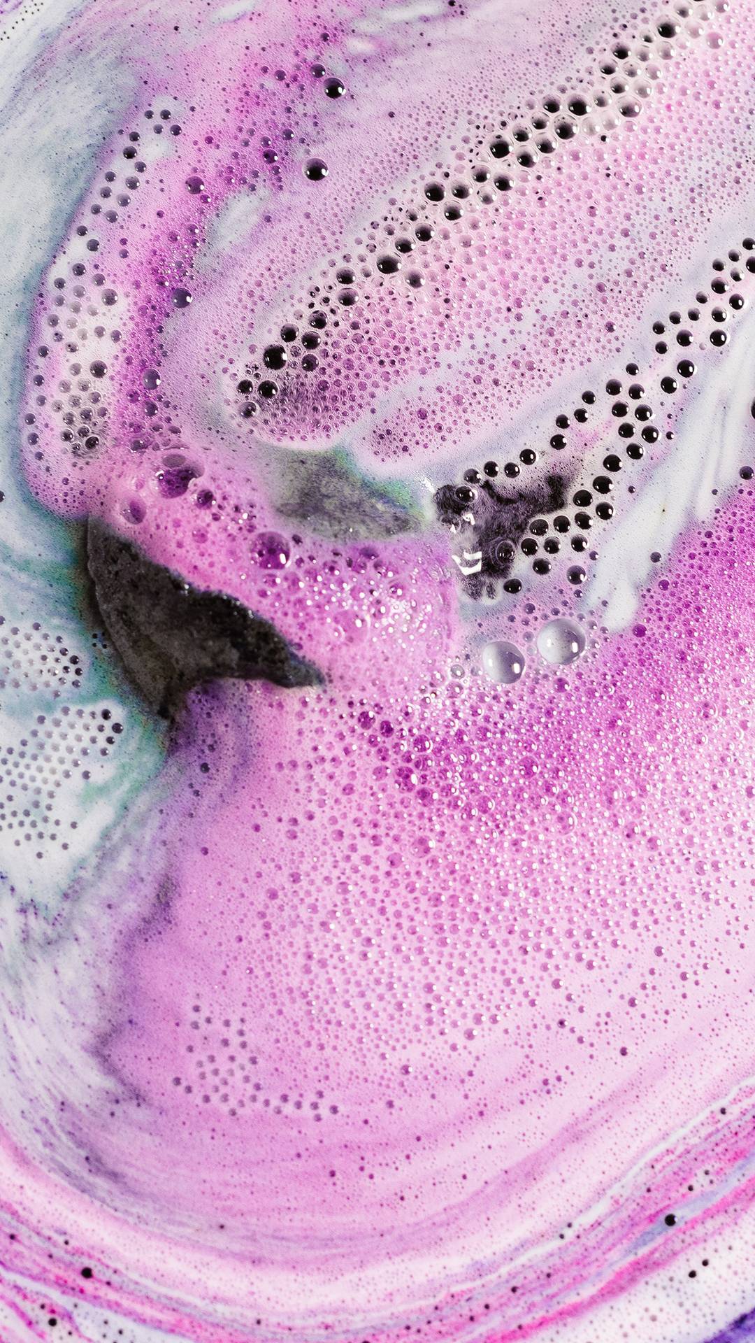The Black Rose bath bomb is slowly dissolving creating a velvety galaxy pattern on the water surface made of pink, blue and purple foam above dark bath water. 