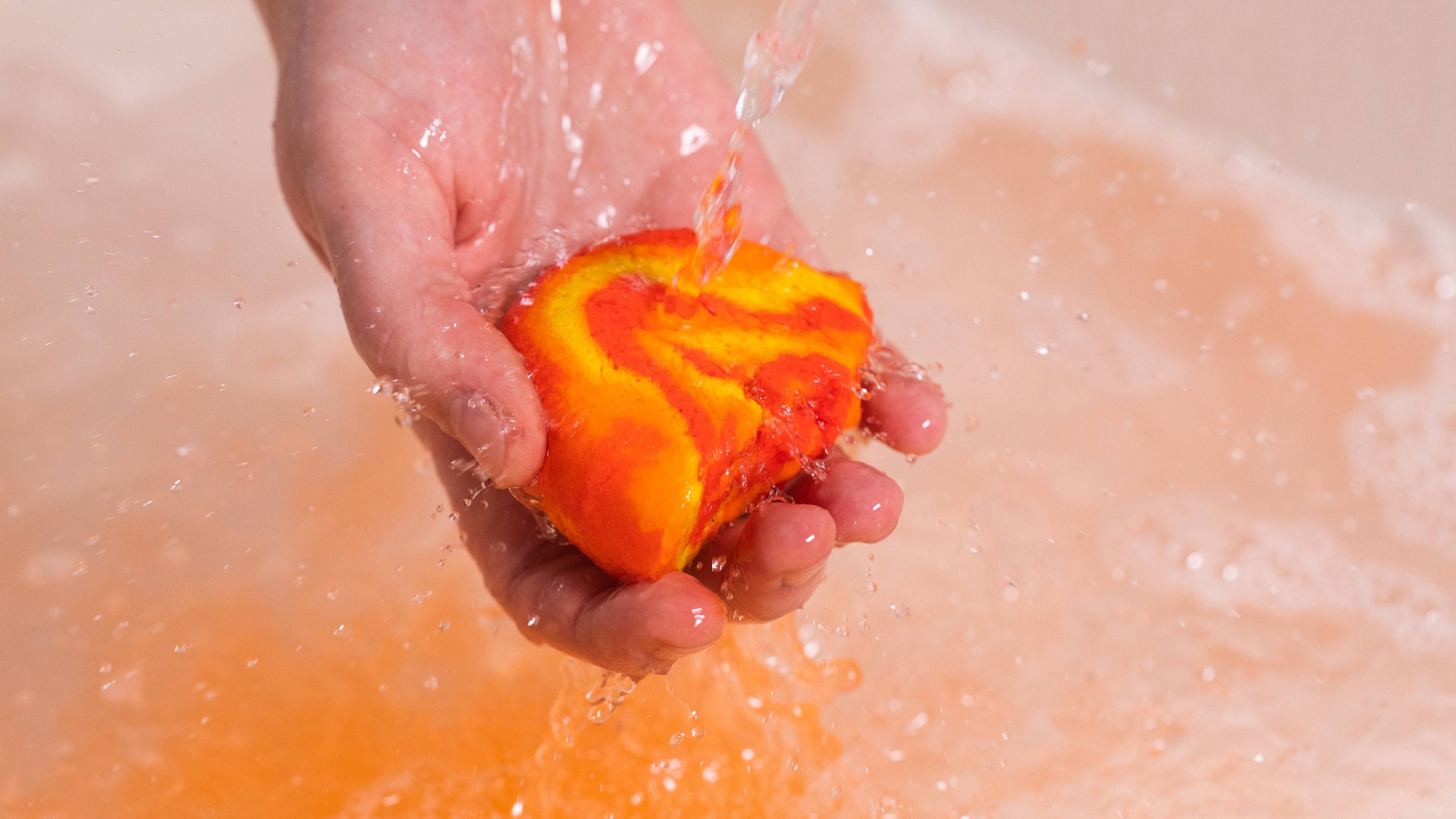The image shows the model's hand under running water as they hold half of the Brightside bubble bar to create a warm orange bath with thick white bubbles below. 