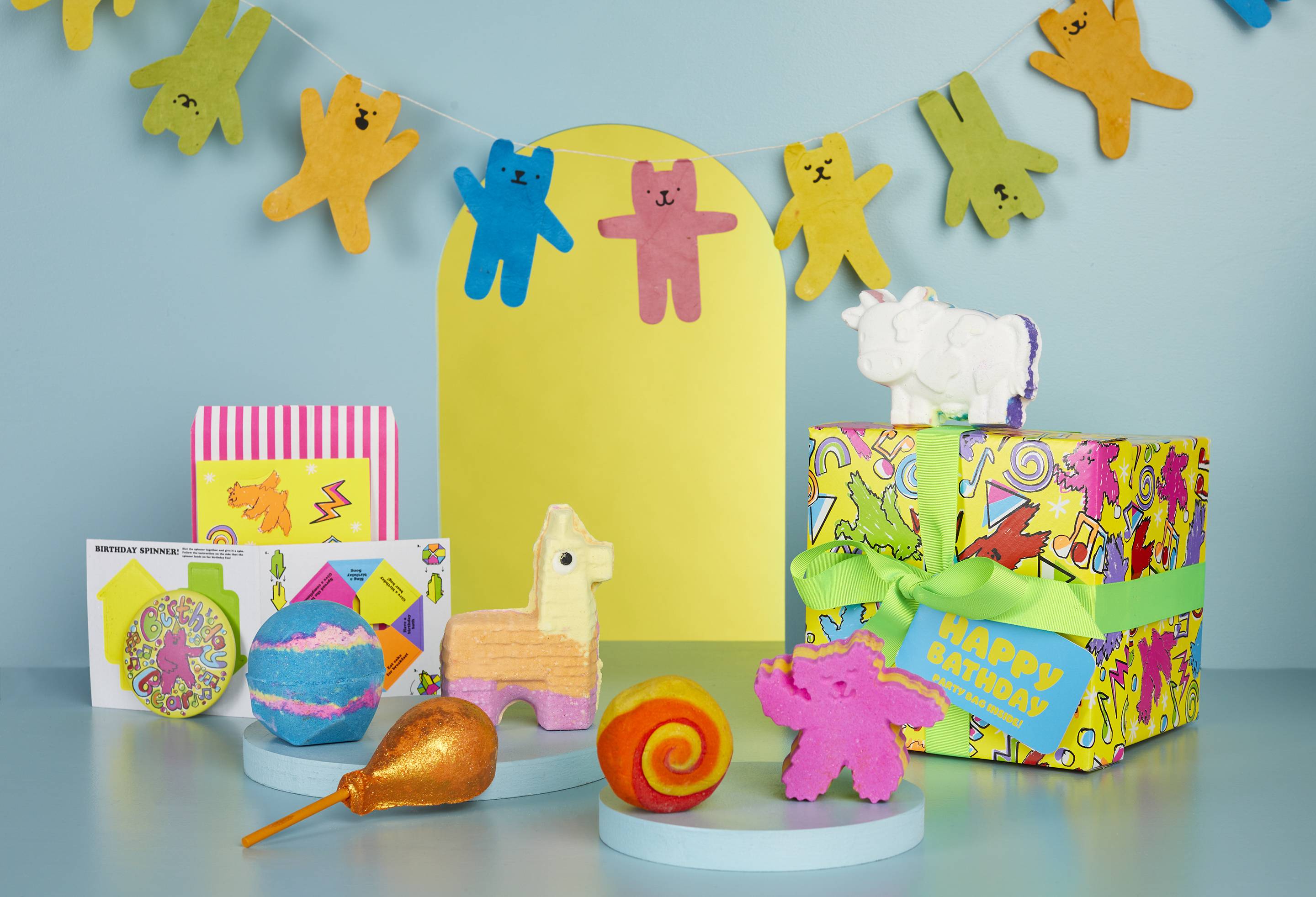 The Happy Bathday gift box, along with the six bath products and extra goodies included, is sat on a pale blue background. There is a yellow arch shape behind it and a banner with different coloured dancing bears attached. 