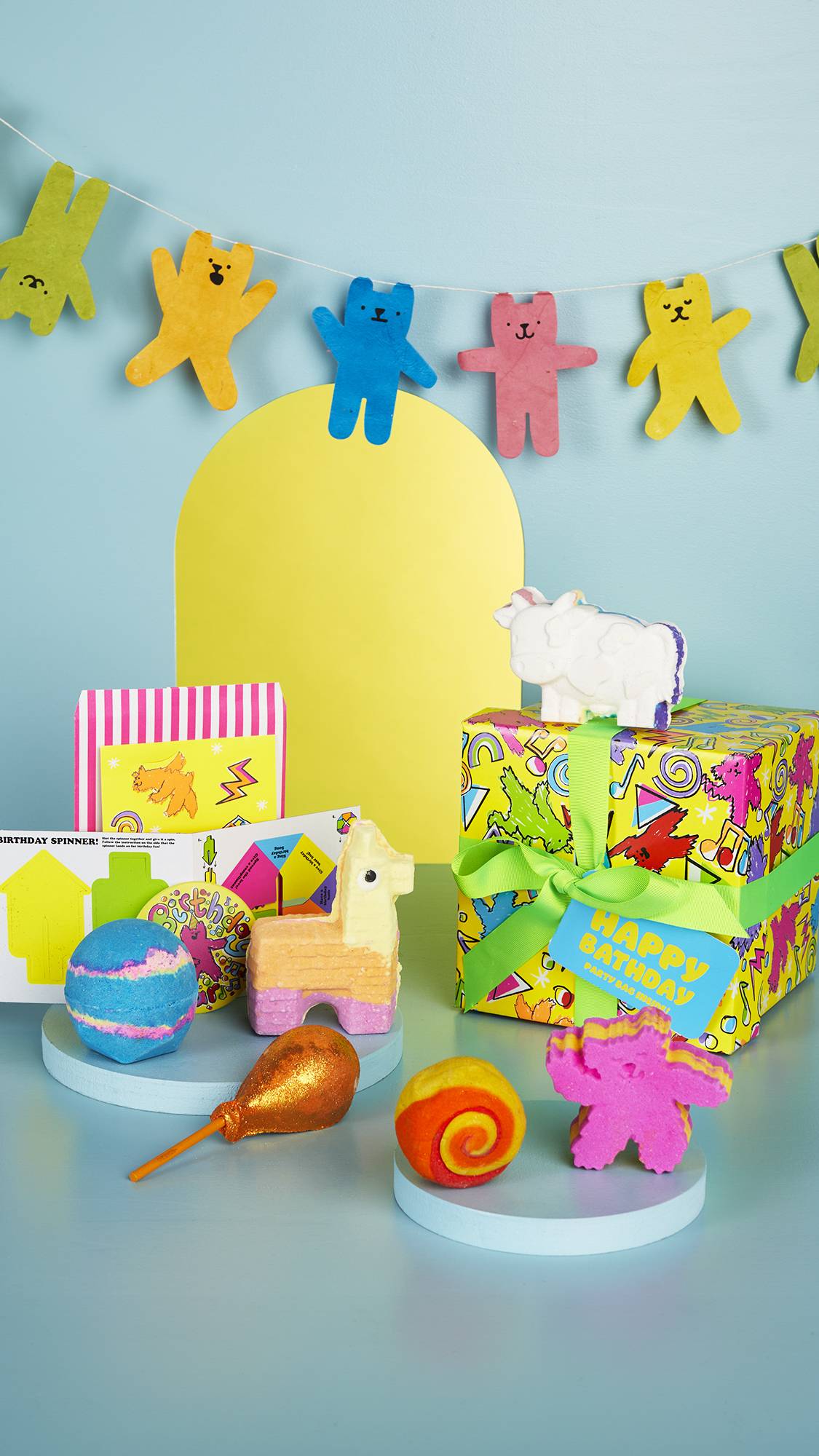 The Happy Bathday gift box, along with the six bath products and extra goodies included is sat on a pale blue background. There is a yellow arch shape behind it and a banner with different coloured dancing bears attached. 