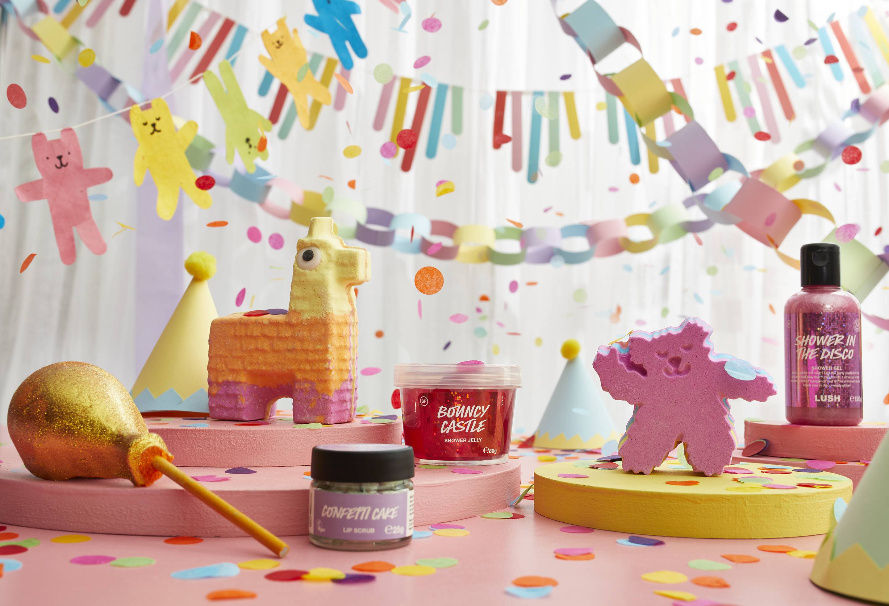 A table is scattered with colourful confetti with the six, Lush party products sitting on circular, flat, wooden wedges with multiple colourful banners, party hats and flying confetti. 