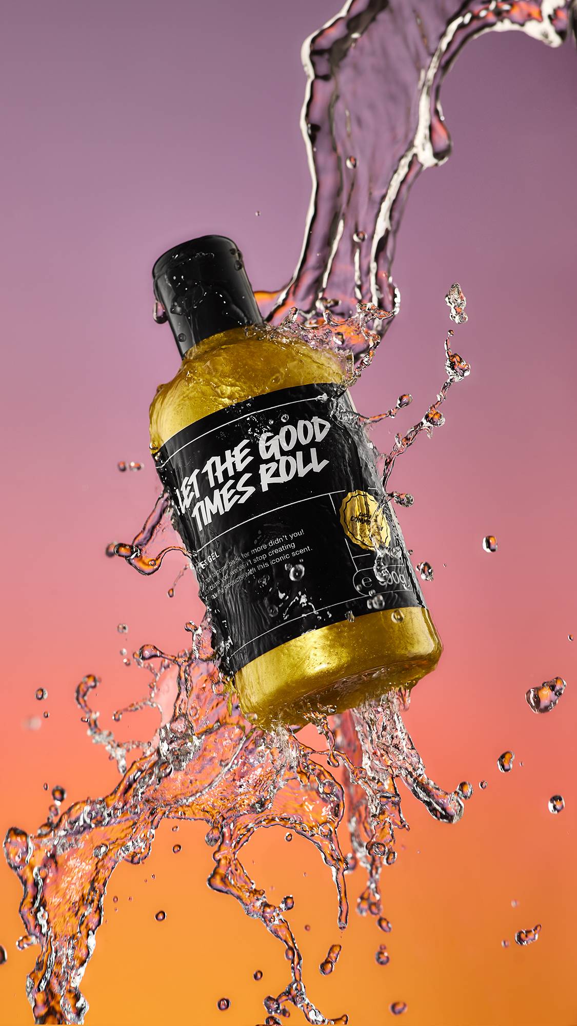 A high-definition action shot of the Let The Good Times Roll shower gel bottle mid-air being splashed by fresh water surrounded by droplets on a blurred purple and orange background.