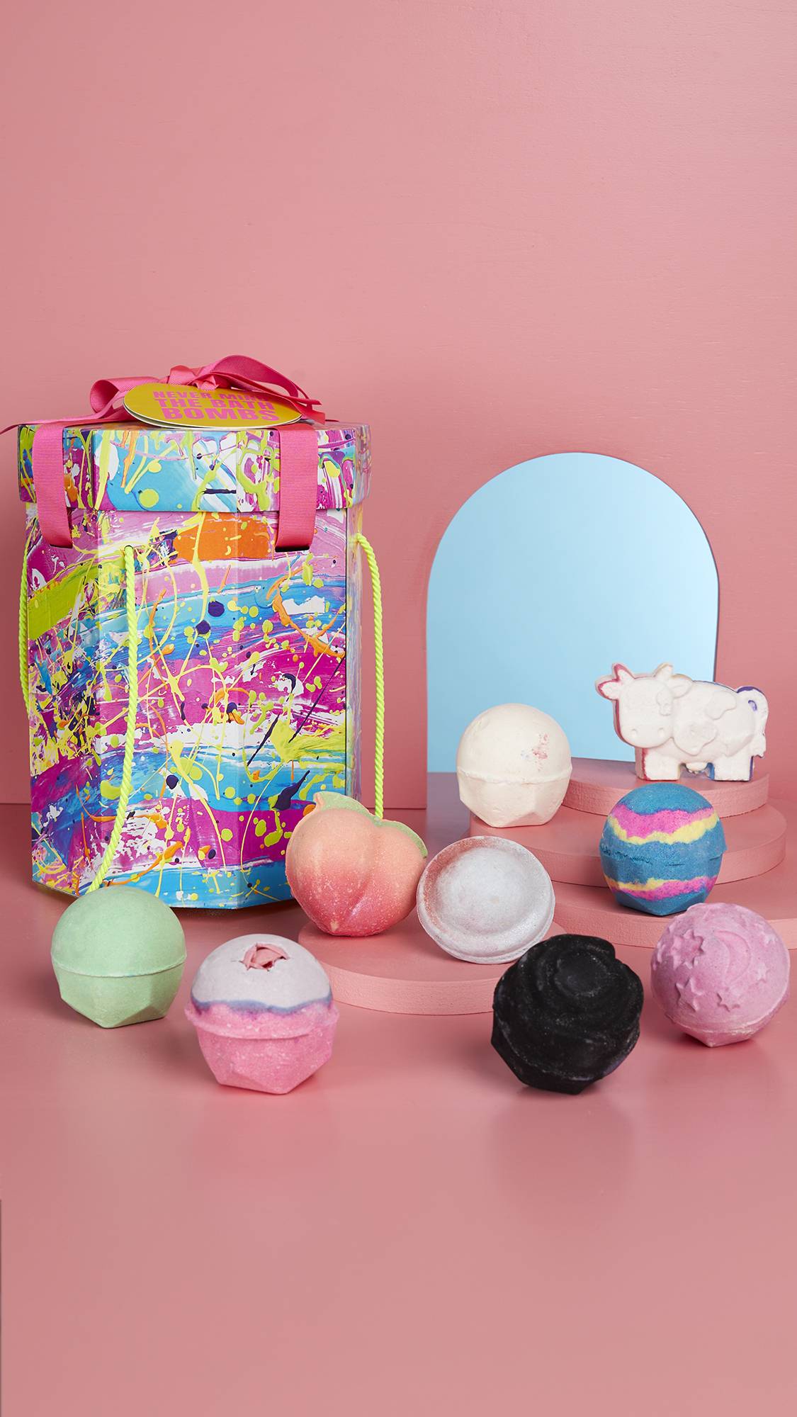 The Never Mind The Bath Bombs gift box is standing to show its full size. It is standing with the nine bath bombs on a pastel pink background. There is an arched mirror behind the products. 