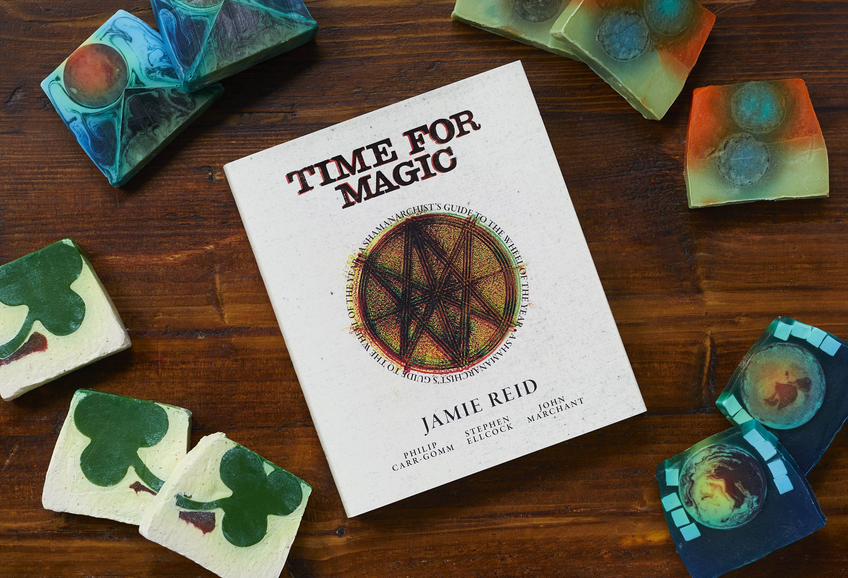 The book "Time For Magic" by Jaime Reid is laid flat on a wooden table surrounded by multiple slices of the four different soap slices designed for Lush with the inspiration of Jaime Reid's art. 